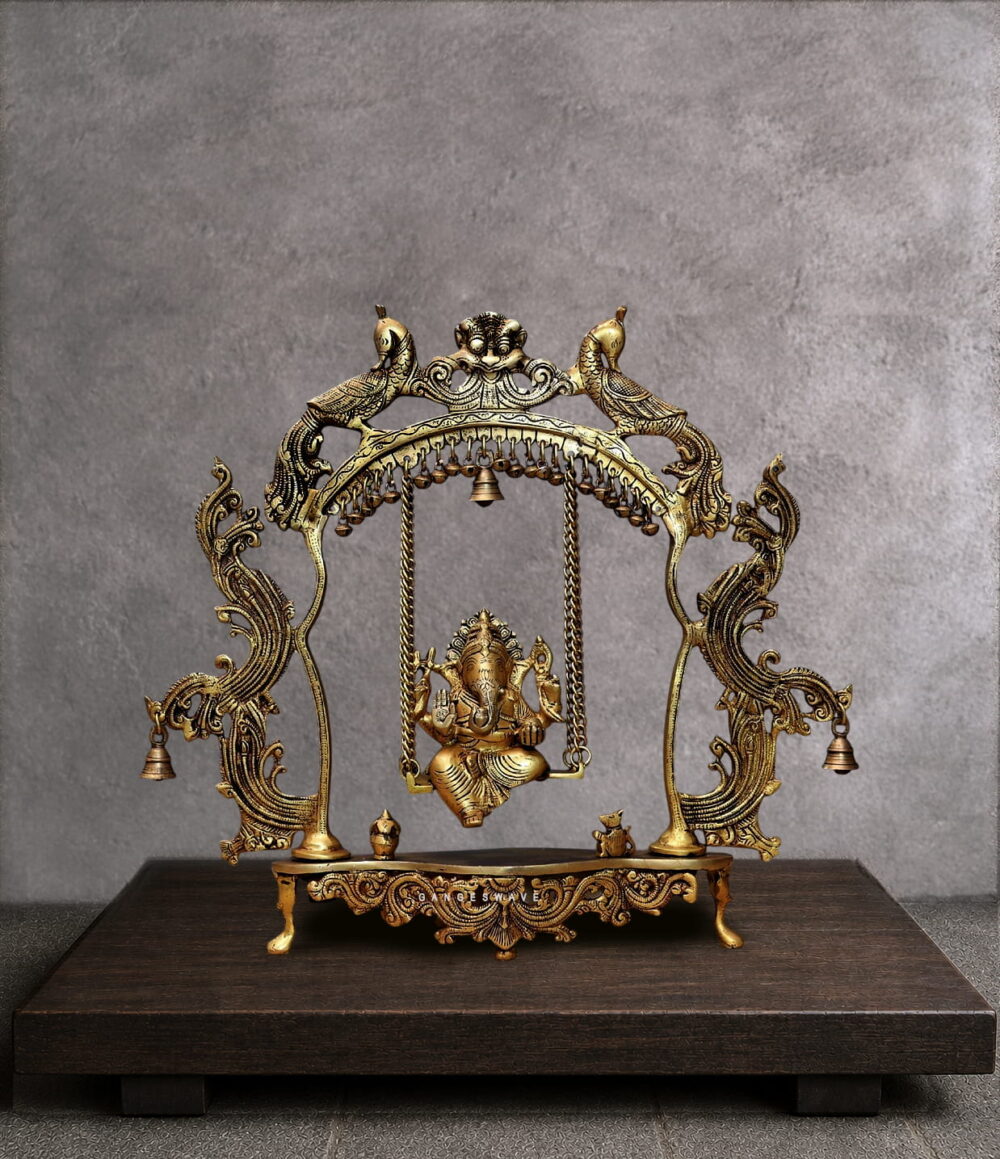 Handcrafted brass Ganesha jhula idol showing Lord Ganpati seated on an ornate swing suspended from a peacock arch with hanging bells. Antique golden finish with intricate filigree details. Perfect for pooja room and spiritual home decor.