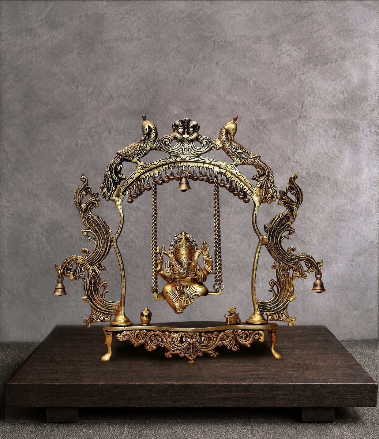 Handcrafted brass Ganesha jhula idol showing Lord Ganpati seated on an ornate swing suspended from a peacock arch with hanging bells. Antique golden finish with intricate filigree details. Perfect for pooja room and spiritual home decor.
