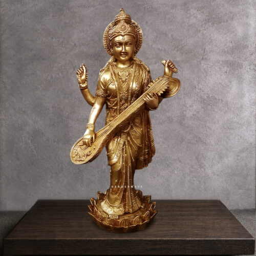 Close-up detail of brass Saraswati idol face showing serene meditative expression, ornate jeweled crown with floral motifs, third eye, and traditional Indian goddess ornamentation. Handcrafted brass deity facial detail.