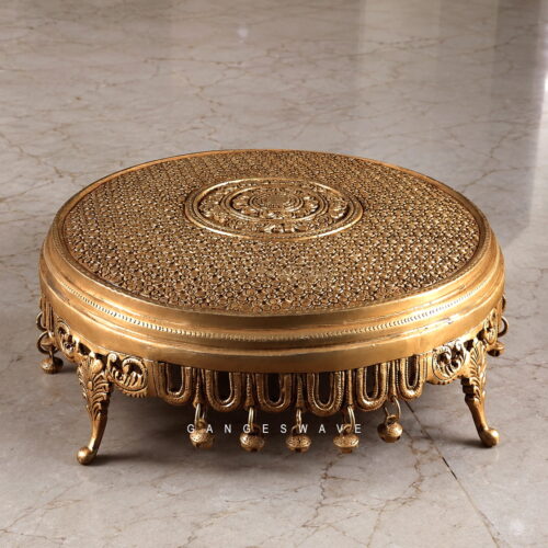 Ornate round antique brass chowki with intricate floral engraving, decorative legs and hanging bells for pooja idols and luxury home décor