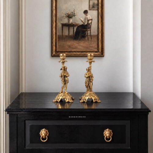 Pair of vintage brass figurine candle holders with ornate bases and golden finish displayed on a dark console.